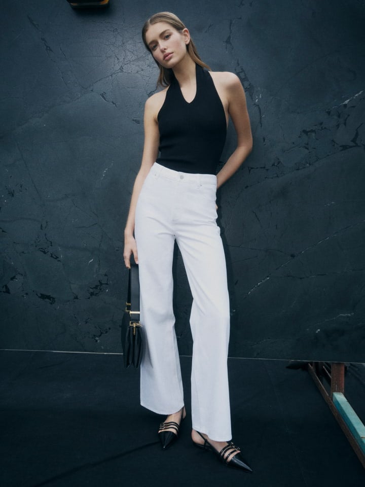 Woman in black halter neck knit, white high rise wid leg jean, black michelle flats and black liv mini leather bag